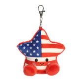 A small red, white, and blue star plush clip-on with pom-pom feet from Palm Pals by Aurora.